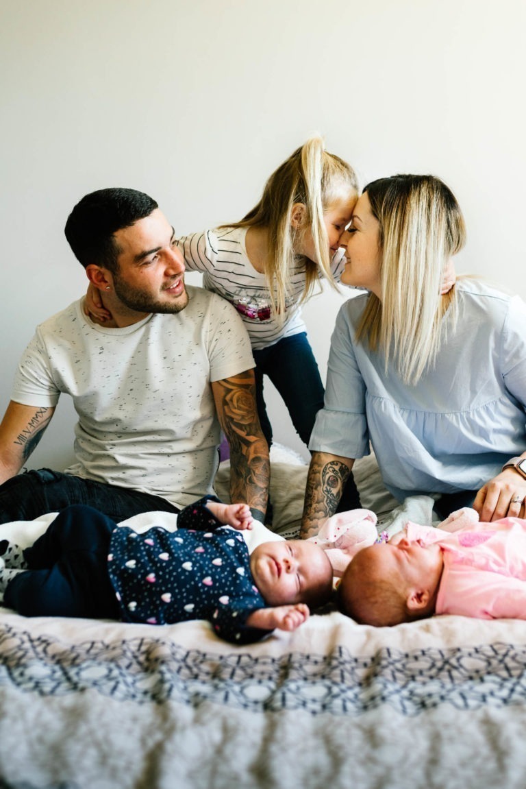 Un père tatoué est assis sur un lit, vêtu d’un t-shirt blanc, et sourit à sa fille blonde qui embrasse sa mère. La mère, également tatouée, porte un chemisier bleu clair. Deux nourrissons sont allongés sur le lit, l’un dans une tenue bleu marine avec des étoiles et l’autre dans une combinaison et un chapeau roses, se reposant paisiblement. C’est comme si leur histoire d’amour avait commencé par un coup de foudre au feu.