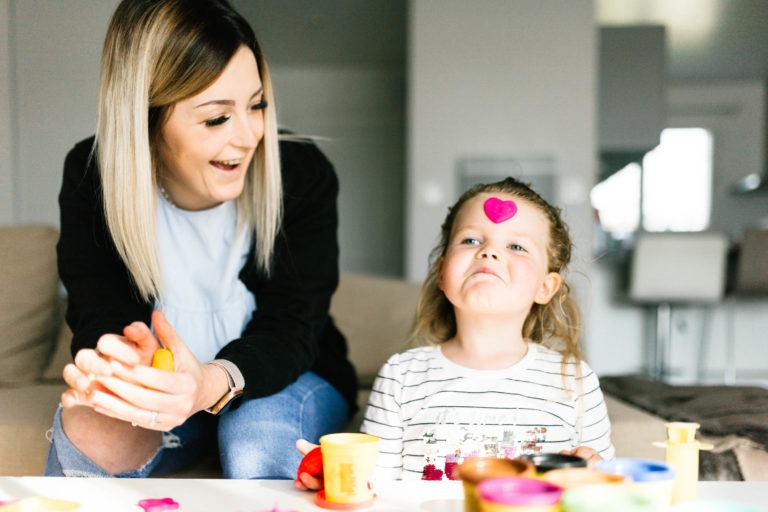 Une femme aux cheveux blonds et un enfant sont assis à une table avec des peintures de différentes couleurs. La femme sourit et roule de l'argile tandis que l'enfant, portant un autocollant en forme de cœur rose sur le front, lève les yeux avec une expression enjouée. La scène à l'intérieur ressemble à un coup de foudre au feu rouge, avec une cuisine moderne visible en arrière-plan.