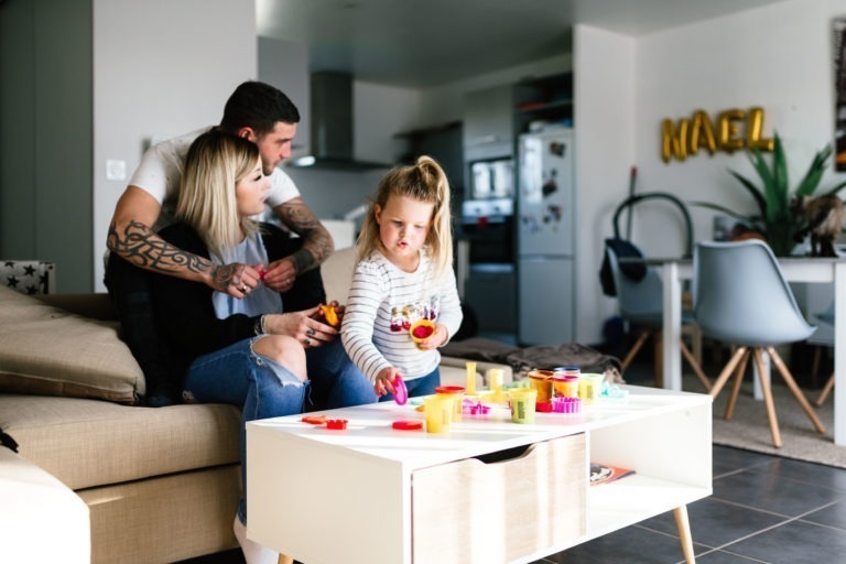 Une famille est assise dans le salon. Un homme, une femme et une jeune fille avec une queue de cheval jouent avec de la pâte à modeler aux couleurs vives devant une table basse blanche. La femme et la fille sont assises sur un canapé beige, tandis que l'homme se tient derrière eux. En arrière-plan, des appareils de cuisine et des ballons « NOEL » sont visibles, une scène de bonheur douillet qui rappelle le coup de foudre au feu rouge.