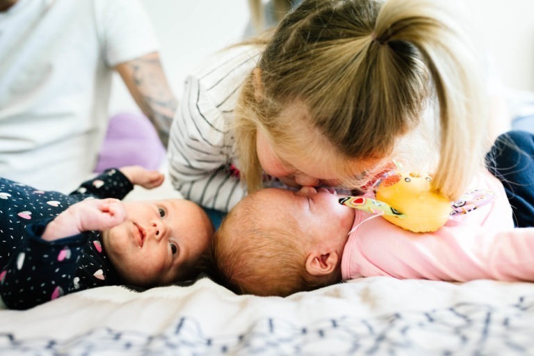 Un jeune enfant avec une queue de cheval haute embrasse doucement le front d'un bébé allongé sur un lit. Un autre bébé, vêtu d'une tenue sombre à motifs colorés, regarde avec curiosité. La scène est chaleureuse et tendre, capturant des moments magiques de complicité presque comme un coup de foudre au feu rouge entre frères et sœurs.