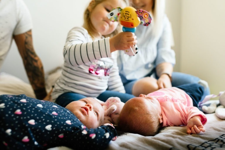 Un jeune enfant tient un jouet coloré au-dessus de deux nourrissons allongés sur un lit. L'un des bébés est vêtu d'une combinaison sombre à motifs de cœurs, et l'autre porte une tenue rose clair. Deux adultes, partiellement visibles, sont assis à l'arrière-plan, en train de discuter avec les enfants dans un cadre intérieur chaleureux, un véritable coup de foudre au feu rouge.