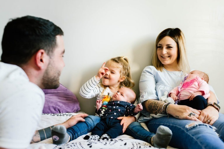 Un homme et une femme, tous deux souriants, sont assis sur un lit avec deux jeunes enfants. La femme, tenant un nouveau-né habillé en rose, est assise à côté d'une fillette portant un haut blanc et un jean bleu qui touche de manière ludique le nez d'un bébé habillé en sombre sur les genoux de l'homme. Cette famille, semble-t-il, vit son propre coup de foudre au feu rouge, un moment de joie inattendu.