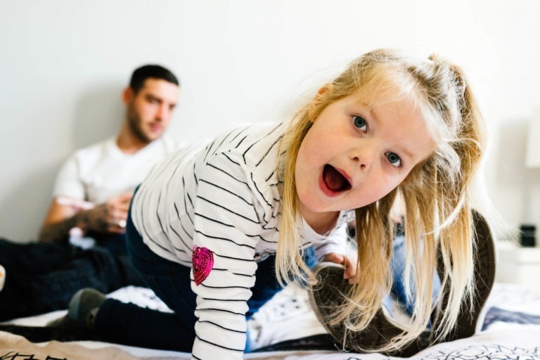 Une jeune fille blonde portant un chemisier rayé avec un patch en forme de cœur rose se penche avec empressement vers la caméra avec une expression large et joyeuse. À l'arrière-plan, un homme, légèrement flou, est assis sur un lit et la regarde nonchalamment. La scène transmet le sentiment ludique et intime du coup de foudre au feu rouge dans ce moment familial.