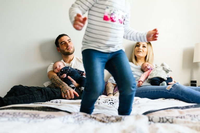 Une famille est assise sur un lit : un homme porte un bébé, une femme berce un autre bébé et un jeune enfant se tient au premier plan, dos à la caméra. L'homme et la femme sourient, créant une atmosphère chaleureuse et joyeuse rappelant le coup de foudre au feu rouge.