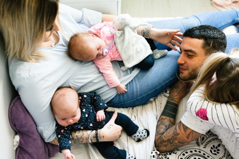 Une famille de quatre personnes se détend. Une femme tient dans ses bras un bébé vêtu d'une tenue rose, tandis qu'un homme tatoué les regarde, rappelant le coup de foudre au feu rouge. Un autre bébé, habillé en bleu foncé, est allongé sur les genoux de la femme. Une jeune fille avec une queue de cheval est également présente, partiellement visible, en train d'interagir avec l'homme.