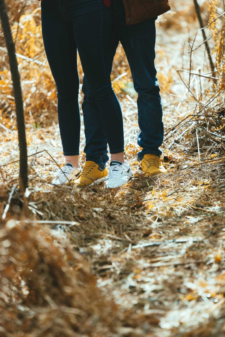 Deux personnes se tiennent côte à côte dans un sentier forestier ensoleillé, portant un jean bleu foncé, lors d'une séance photo en amoureux à Bordeaux. La personne de gauche porte des baskets blanches, tandis que celle de droite en porte des jaunes vifs. Le sol est couvert de feuilles et de brindilles séchées, ainsi que de hautes herbes.