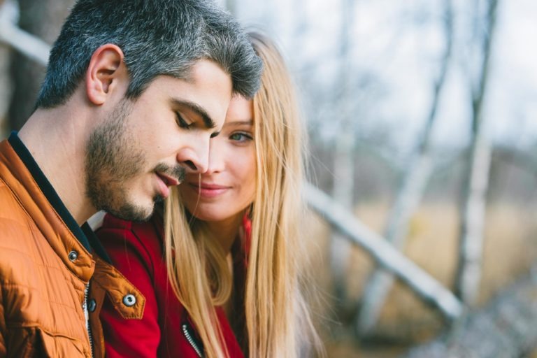 Un couple partage un moment de tendresse en extérieur lors d'une séance photo en amoureux à Bordeaux. L'homme aux cheveux gris courts et à la barbe porte une veste marron, se penchant doucement vers la femme. La femme aux longs cheveux blonds lui sourit doucement, portant une veste rouge. L'arrière-plan est un décor naturel flou avec des arbres.