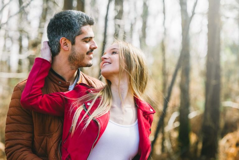 Un homme aux cheveux courts et noirs, vêtu d'une veste marron, se tient enlacé avec une femme blonde portant une veste rouge et un haut blanc, tandis qu'ils se sourient et se regardent. Ils sont à l'extérieur, dans une zone boisée et ensoleillée avec des arbres nus en arrière-plan, capturant leur séance photo en amoureux à Bordeaux.