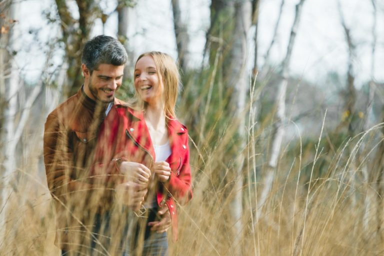 Un couple se tient près l'un de l'autre dans un champ herbeux, souriant et riant. L'homme, vêtu d'une veste marron, se tient derrière la femme portant une veste rouge. De hautes herbes sèches les entourent et des arbres aux troncs nus peuvent être vus en arrière-plan. Cette photo de séance en amoureux à Bordeaux capture leur atmosphère joyeuse et légère.