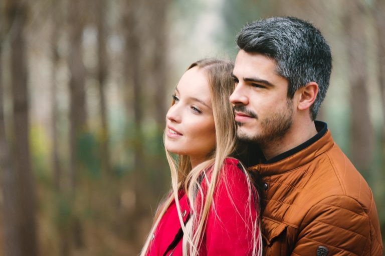 Un couple se tient près l'un de l'autre, l'homme enlaçant la femme par derrière. Tous deux semblent regarder au loin avec des expressions satisfaites. La femme a de longs cheveux blonds et porte une veste rouge, tandis que l'homme a des cheveux noirs courts avec des mèches grises et porte une veste marron. Ils sont entourés d'arbres flous dans un décor forestier calme, capturant leur séance photo en amoureux à Bordeaux.