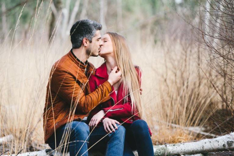 Un couple est assis sur un tronc d'arbre tombé dans une forêt près de Bordeaux, et s'embrasse. L'homme porte une veste marron et un jean, tenant doucement le menton de la femme. La femme, vêtue d'une veste rouge et d'un jean avec de longs cheveux blonds, se penche vers elle. De hautes herbes et des arbres flous les entourent, mettant en valeur leur séance photo sereine en amoureux.