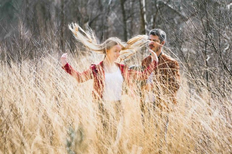 Une femme et un homme se tiennent dans un champ ensoleillé d'herbes hautes et sèches pendant leur séance photo en amoureux à Bordeaux. La femme, aux longs cheveux blonds, tourne sur elle-même, faisant tournoyer ses cheveux. Tous deux sont habillés de vêtements décontractés. Des arbres nus à l'arrière-plan indiquent un décor d'automne ou de début de printemps, créant une atmosphère légère et joyeuse.