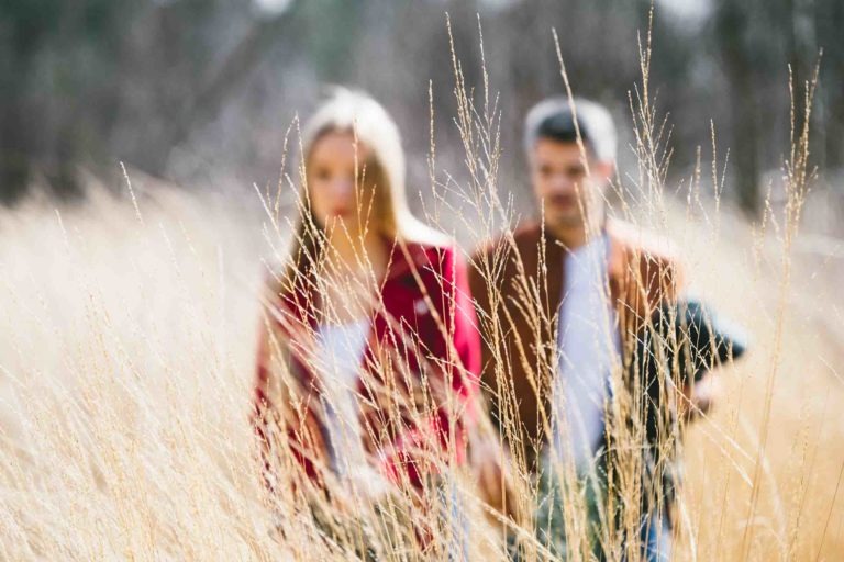 Un homme et une femme marchent dans un champ ensoleillé d'herbes hautes, capturés dans une séance photo onirique et floue en amoureux à Bordeaux. La femme en veste rouge et l'homme en veste marron tenant une chemise à carreaux se fondent dans l'herbe dorée, avec des arbres flous peignant l'arrière-plan.