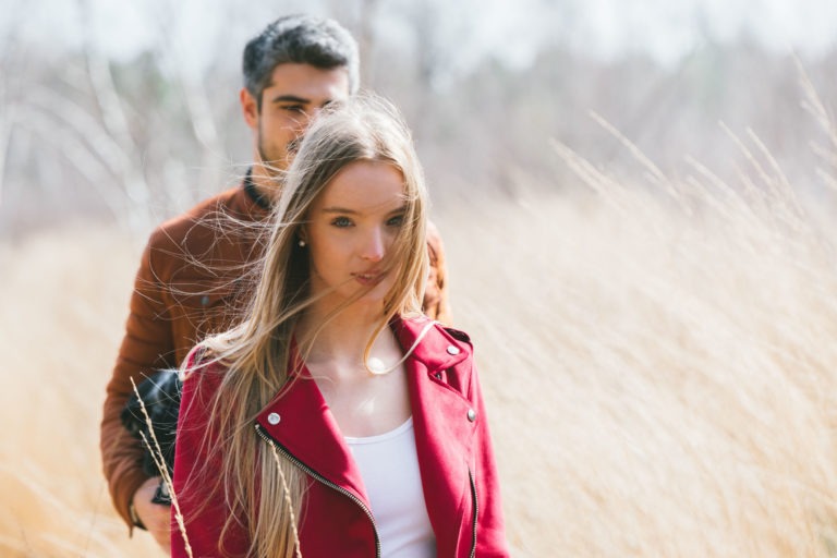 Une jeune femme aux longs cheveux blonds, vêtue d'une veste rouge, marche dans les hautes herbes sèches. Derrière elle se trouve un homme aux cheveux courts et foncés, vêtu d'une veste marron. Tous deux ont une expression pensive sur cette photo de séance en amoureux à Bordeaux, sur fond d'arbres flous et nus.