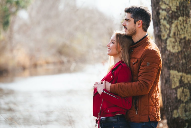 Un couple se tient près d'un arbre au bord d'un plan d'eau. La femme porte une veste rouge et sourit avec contentement tandis que l'homme, vêtu d'une veste matelassée marron, se tient derrière elle, regardant au loin. Cette séance photo sereine en amoureux à Bordeaux présente des arbres légèrement flous et de l'eau en arrière-plan.