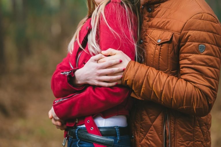 Gros plan d'un couple en train de s'embrasser en plein air. La personne à gauche, vêtue d'une veste rouge, est serrée dans ses bras par derrière par la personne à droite, vêtue d'une veste matelassée orange-marron. Les visages du couple ne sont pas visibles, l'accent étant mis sur leur proximité et leur étreinte lors d'une séance photo en amoureux à Bordeaux.