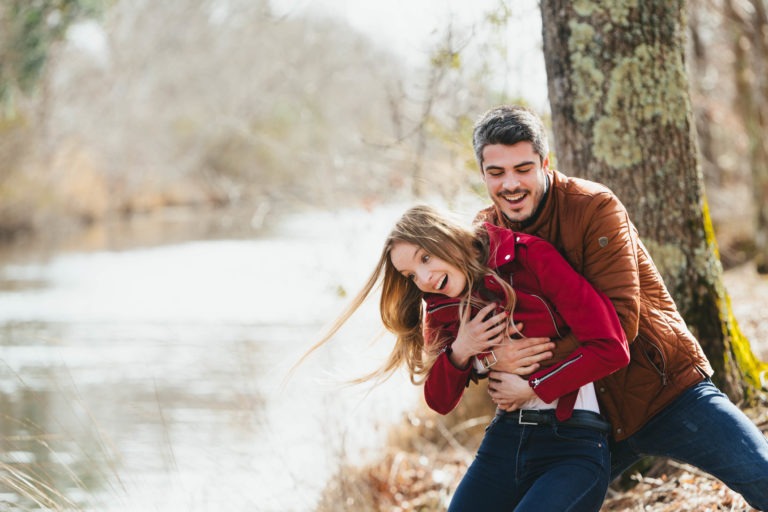 Un jeune couple rit debout près d'un arbre au bord d'une rivière, dans ce qui semble être une séance photo joyeuse en amoureux à Bordeaux. L'homme, aux cheveux noirs courts et à la barbe, portant une veste marron, embrasse la femme par derrière. Elle, aux longs cheveux blonds et à la veste rouge, se penche en avant de manière ludique dans ce magnifique décor extérieur.