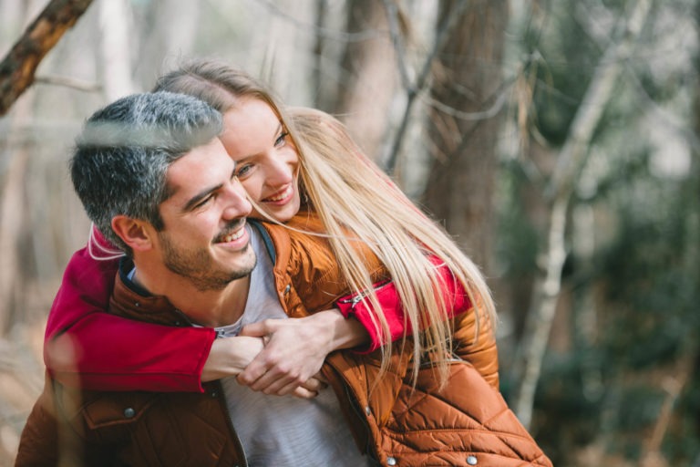 Un homme souriant aux cheveux courts et grisonnants et à la barbe porte sur son dos une femme souriante aux longs cheveux blonds. Ils portent des vestes chaudes dans ce qui ressemble à une séance photo en amoureux à Bordeaux, avec des arbres flous en arrière-plan suggérant qu'il fait froid.