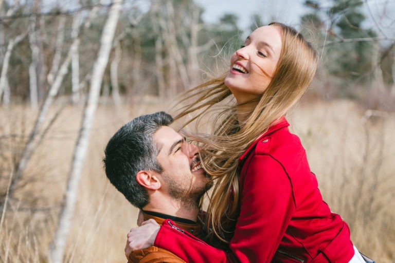 Une femme souriante aux longs cheveux blonds et à la veste rouge est joyeusement soulevée du sol par un homme aux cheveux noirs courts et à la barbe portant une veste marron. Dans cette scène de séance photo en amoureux à Bordeaux, ils se tiennent dans un champ d'herbe sèche avec des arbres sans feuilles, et la femme rit la tête renversée, créant une scène de bonheur et d'affection.