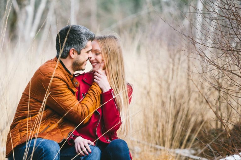 Un couple est assis côte à côte dans un espace naturel herbeux entouré d'herbe sèche marron clair et de branches nues pendant leur séance photo en amoureux à Bordeaux. L'homme, vêtu d'une veste orange, regarde et sourit à la femme portant une veste rouge alors qu'ils partagent un moment de tendresse.