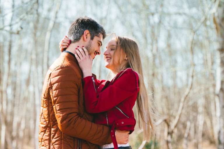 Un couple se tient debout, étroitement enlacé, dans un espace boisé en plein air. L'homme, vêtu d'une veste marron, et la femme, vêtue d'une veste rouge, se regardent amoureusement, leurs visages proches, tous deux souriants. L'arrière-plan présente des arbres sans feuilles, suggérant le début du printemps ou la fin de l'automne - une belle séance photo en amoureux à Bordeaux.