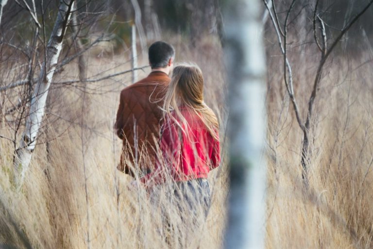 Un homme et une femme marchent côte à côte sur un étroit sentier bordé d'herbes hautes et sèches. On les voit de dos, l'homme portant une veste marron et la femme une veste rouge. Des branches d'arbres nues se courbent au-dessus du sentier, créant un effet de tunnel. La scène respire l'ambiance calme et paisible d'une séance photo en amoureux à Bordeaux.