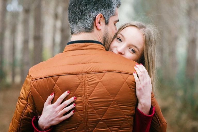 Une femme aux cheveux blonds et aux ongles vernis rouge serre un homme par derrière. L'homme a les cheveux courts et grisâtres et porte une veste matelassée marron. Ils se tiennent au milieu d'une forêt aux arbres nus. La femme sourit tout en posant sa tête sur l'épaule de l'homme, transmettant un sentiment de chaleur et d'affection dans cette séance photo en amoureux à Bordeaux.