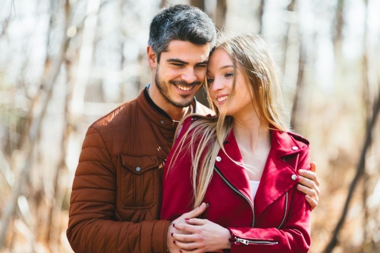 Un homme aux cheveux courts et foncés et à la barbe enlace une femme aux cheveux longs et blonds. Tous deux sourient. Il porte une veste marron et elle une veste rouge. Ils sont en extérieur à Bordeaux, avec des arbres flous et la lumière du soleil filtrant sur cette photo de séance en amoureux.