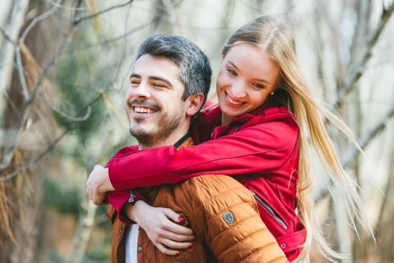 Un couple heureux dehors, avec une femme en veste rouge qui porte un homme en veste marron sur son dos. Ils ont tous les deux le sourire aux lèvres. L'arrière-plan montre des arbres flous, suggérant un décor de forêt ou de parc à Bordeaux. L'atmosphère est lumineuse et joyeuse, capturant cette joyeuse séance photo en amoureux.