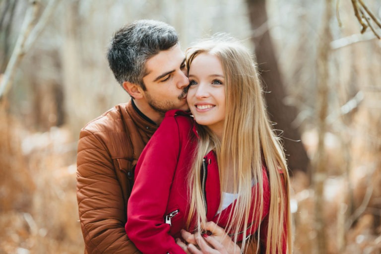 Un homme aux cheveux noirs et à la barbe, portant une veste marron, enlace une femme souriante aux longs cheveux blonds qui porte une veste rouge. Ils sont dehors dans une forêt avec des arbres flous en arrière-plan pendant leur séance photo en amoureux à Bordeaux. L'homme embrasse la femme sur la joue, et tous deux ont l'air joyeux et satisfaits.