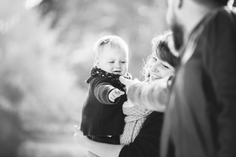 Une image en niveaux de gris montre un jeune enfant porté par une femme. L'enfant, vêtu d'un pull tricoté, tend le bras vers un homme partiellement visible à droite. La femme, souriante et portant un foulard, regarde l'enfant avec amour. L'arrière-plan flou suggère un décor extérieur d'épanouissement familial.