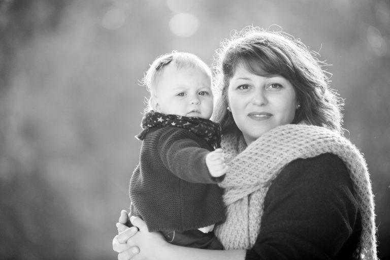 Une photo en noir et blanc montre un adulte tenant un bébé dans ses bras. L'adulte, vêtu d'une écharpe et d'un pull tissés, regarde la caméra avec une expression douce de famille épanouissement. Le bébé, également tourné vers la caméra, porte un bandeau et un pull tricoté, étendant son bras droit légèrement vers l'avant. L'arrière-plan est flou.