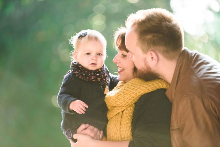 Un couple souriant tient un bébé dehors sous un soleil éclatant. La femme, portant un foulard jaune, soutient le bébé tandis que l'homme se penche pour l'embrasser sur la joue. Le bébé, vêtu d'un pull sombre et d'un foulard à motifs, regarde directement la caméra avec une expression neutre. Un feuillage vert fleurit derrière cette scène d'épanouissement familial.