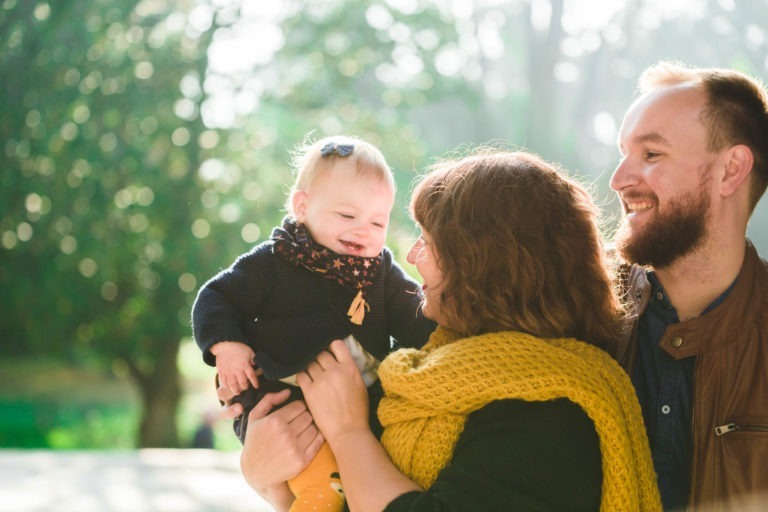 Une famille heureuse est dehors par une journée ensoleillée. Une femme aux cheveux bruns ondulés jusqu'aux épaules porte un foulard jaune vif, tenant un bébé souriant vêtu d'une tenue sombre et d'un foulard à motifs d'étoiles. Un homme barbu se tient à côté d'eux, regardant le bébé avec une expression joyeuse. Épanouissement parmi les arbres en arrière-plan.