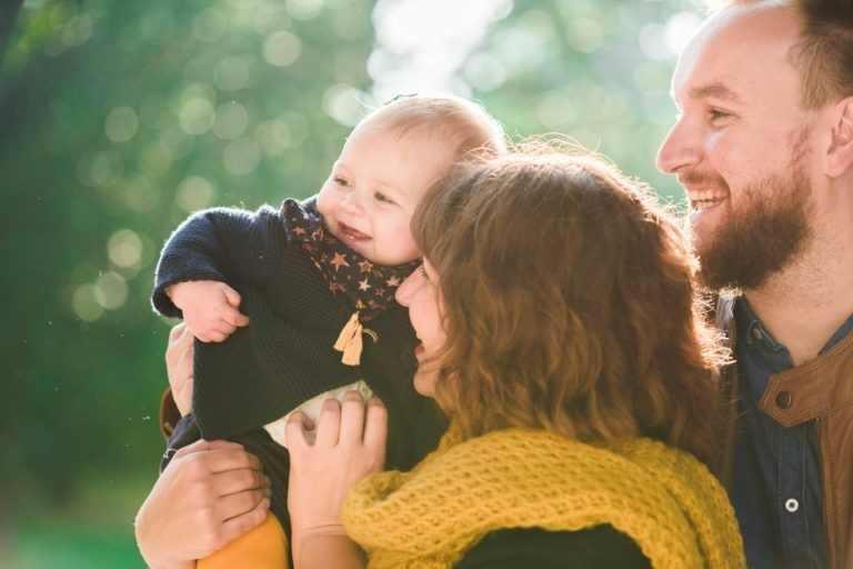 Un moment de joie en famille en plein air. Un homme barbu et une femme aux cheveux bouclés, tous deux souriants, tiennent un bébé rieur vêtu d'une tenue bleu marine. Le bébé a l'air ravi, portant un foulard à motifs étoilés. La femme a un foulard jaune autour du cou. La lumière du soleil filtre à travers un fond vert luxuriant, reflétant leur épanouissement familial.