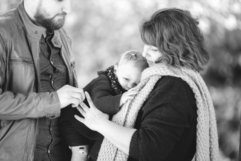 Une photo en noir et blanc montre un homme barbu portant une veste en cuir debout à côté d'une femme portant une écharpe tricotée, tenant un bébé. Le bébé porte un nœud et se blottit contre la femme, tandis que l'homme pose doucement sa main sur le dos du bébé. La scène respire l'épanouissement familial, chaleureux et tendre.