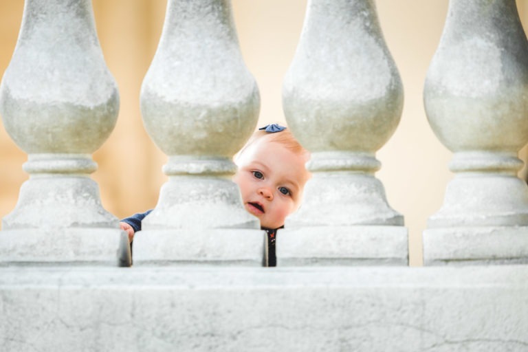 Un bébé jette un œil à travers les interstices entre les larges balustres en pierre. La tête du bébé est légèrement inclinée, avec une expression curieuse sur le visage. Les balustres sont régulièrement espacés, créant une barrière symétrique qui incarne le sentiment d'épanouissement familial dans son cadre serein.