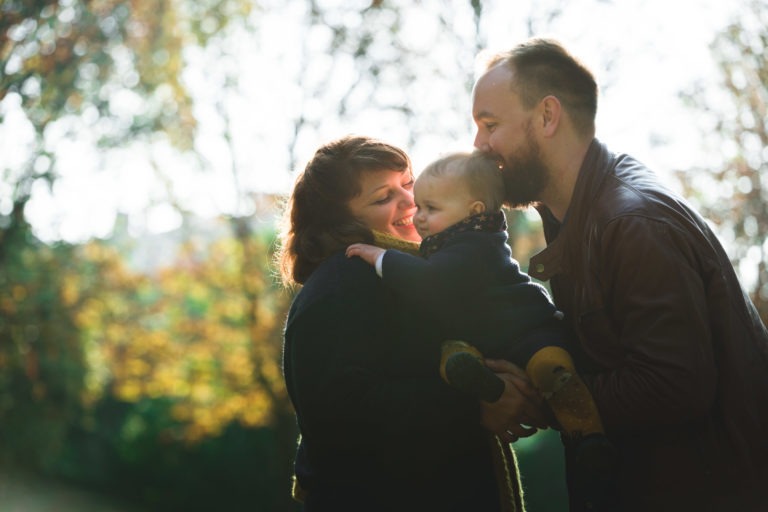 Une famille de trois personnes se tient ensemble dans un parc, rayonnant d'épanouissement. La mère, à gauche, sourit et tient un bébé au centre. Le père, à droite, se penche pour embrasser la tête du bébé. L'arrière-plan légèrement flou révèle les arbres et la lumière du soleil tandis qu'ils profitent de leur tenue chaude.