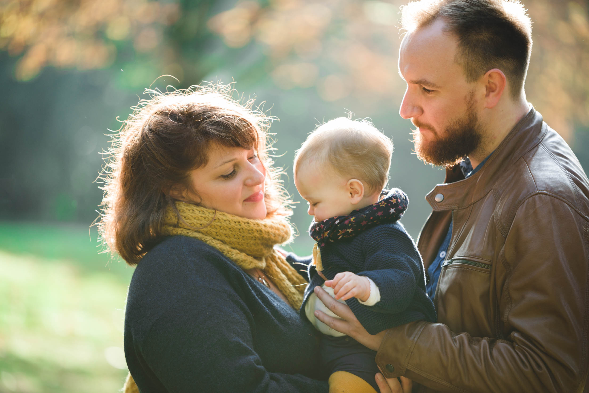 Une mère, un père et leur bébé sont dehors dans un parc sous le soleil. La mère, vêtue d'une écharpe jaune et d'un manteau noir, sourit affectueusement au bébé. Le père, vêtu d'une veste marron, regarde tendrement leur bébé épanoui emmitouflé dans un manteau et une écharpe. L'herbe verte et les arbres sont flous en arrière-plan.