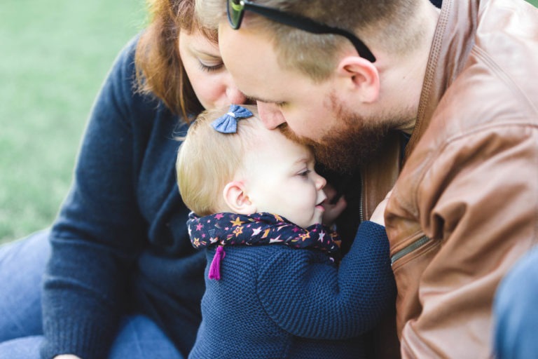 Un homme avec une barbe et des lunettes de soleil sur la tête embrasse amoureusement le front d'un bébé. Le bébé, vêtu d'un pull sombre avec une écharpe colorée et un nœud dans les cheveux, s'appuie contre lui. Une femme aux cheveux bruns, également vêtue d'un pull sombre, observe tendrement le bébé. Ils sont dehors sur l'herbe, rayonnant de famille épanouissement.