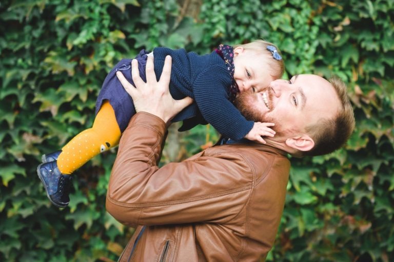 Un homme barbu, vêtu d'une veste en cuir marron, tient un bébé dans ses bras et le soulève vers son visage. Le bébé, vêtu d'une tenue bleu marine et de collants orange vif, presse affectueusement sa joue contre celle de l'homme. Ils se tiennent devant un mur luxuriant recouvert de lierre, incarnant la joie de l'épanouissement familial avec leurs sourires heureux.