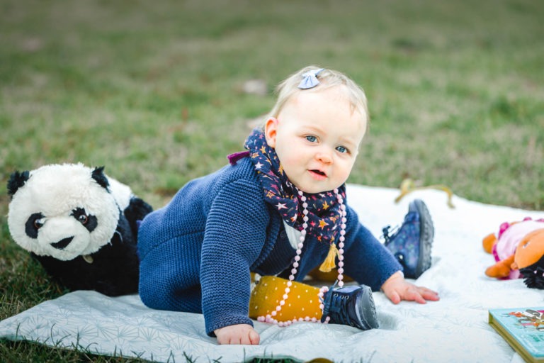 Un bébé est assis sur une couverture de couleur claire sur une pelouse herbeuse, incarnant l'essence de la famille épanouissement. Le bébé porte un pull bleu, une écharpe bleu marine avec des étoiles colorées, un pantalon bleu et des chaussures bleu marine. Un panda en peluche est à côté du bébé à gauche, entouré d'objets éparpillés comme un livre et des jouets.