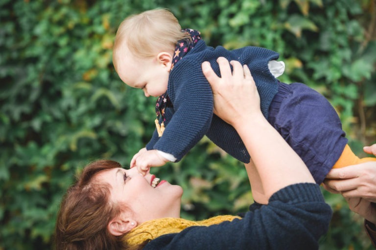 Une personne aux cheveux châtains, coiffée d'un foulard jaune, soulève dans les airs un bébé souriant. Le bébé, vêtu d'un pull bleu foncé et d'un short assorti avec un foulard à motifs d'étoiles, touche le nez de la personne. Dans ce moment d'épanouissement familial, un fond de feuilles vertes est visible.