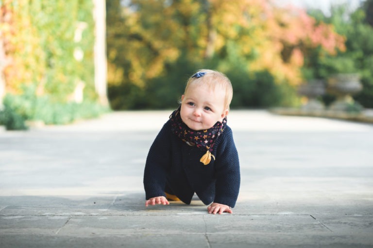 Un jeune bébé portant un pull sombre et une écharpe colorée rampe sur un chemin pavé. Le bébé a les cheveux clairs et un petit nœud attaché près du sommet de la tête, incarnant l'essence de la famille épanouissement. L'arrière-plan présente de la verdure et des arbres aux feuilles d'automne vibrantes dans des tons de vert, de jaune et d'orange.