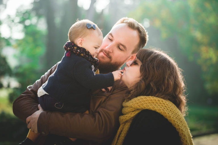 Une famille se tient dehors avec une végétation luxuriante en arrière-plan. Un homme barbu portant une veste en cuir tient un bébé portant un nœud et une écharpe à motifs. Une femme portant une écharpe jaune et un manteau sombre touche doucement la main du bébé, tandis que l'homme et le bébé se regardent avec amour, montrant leur épanouissement.