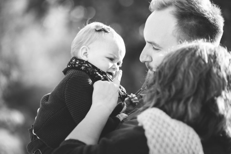 Une photo en noir et blanc montre un bébé tenu par un homme adulte. Le bébé touche le visage de l'homme tandis qu'un autre adulte se tient à proximité. Le bébé porte un nœud dans les cheveux et un pull tricoté, l'homme a une barbe et le deuxième adulte a les cheveux mi-longs et une écharpe texturée, capturant un moment d'épanouissement familial.