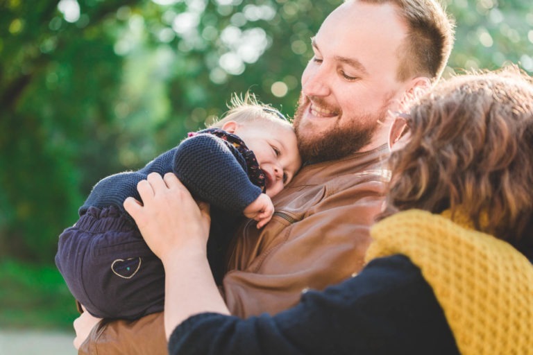 Un homme barbu, vêtu d'une veste marron, tient contre sa poitrine un bébé endormi, vêtu d'une tenue bleu marine. Une femme aux cheveux bouclés, coiffée d'un foulard jaune moutarde, regarde affectueusement le bébé. L'arrière-plan est un décor extérieur vert flou et ensoleillé qui respire la famille épanouissement.