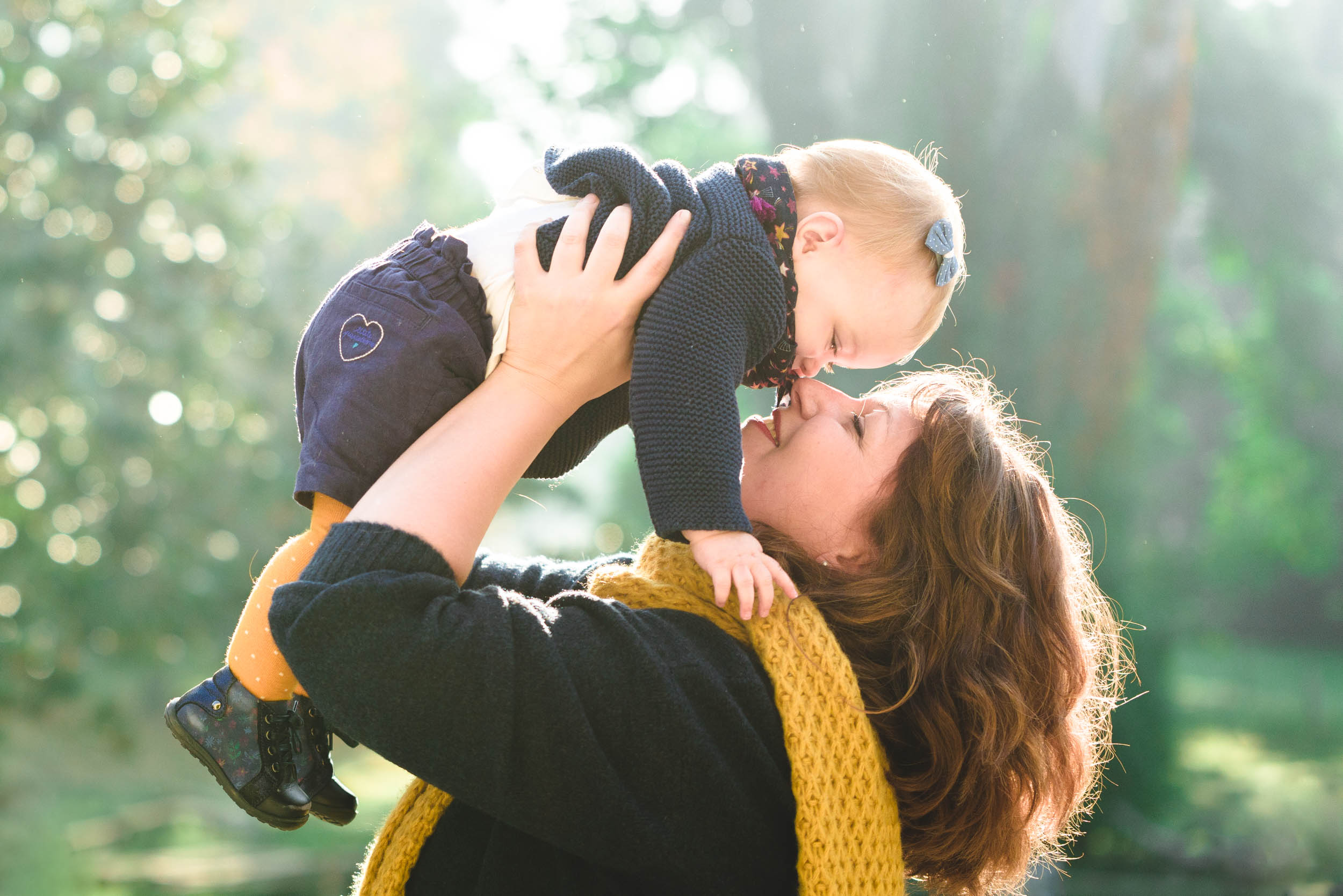 Une femme soulève joyeusement un bébé dans les airs, leurs nez se touchant affectueusement. La femme porte un foulard jaune, tandis que le bébé, orné d'un petit nœud bleu dans les cheveux, porte un pull foncé et un pantalon à motif de cœur. Les arbres ensoleillés en arrière-plan créent une scène chaleureuse et affectueuse d'épanouissement familial.