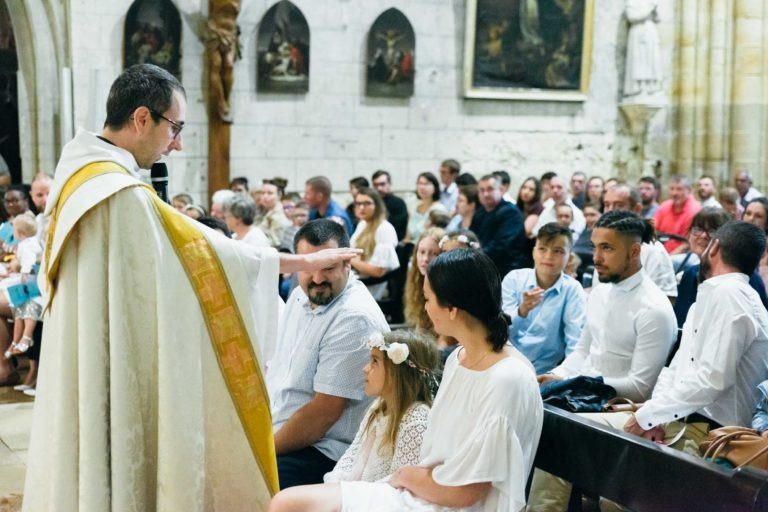 Une photographie de baptême montre un prêtre en robe blanche et or s'adressant à une femme et une jeune fille portant une couronne de fleurs devant une congrégation attentive. L'église, avec ses murs de pierre, ses peintures religieuses et ses statues, crée un décor serein pour ce beau moment.