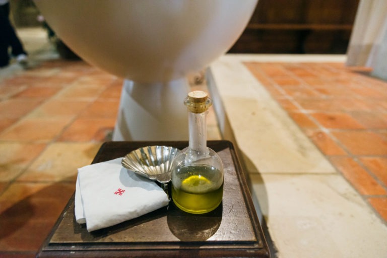 Une petite bouteille remplie d'un liquide vert et fermée par un bouchon de liège est posée sur un support en bois foncé. À côté, un tissu blanc plié sur lequel est brodée une croix rouge. Un plat en argent en forme de coquillage ajoute une touche élégante. L'arrière-plan présente un sol carrelé et des aperçus de structures blanches et en bois, rappelant les objets vus dans les décors de photographes de baptême.